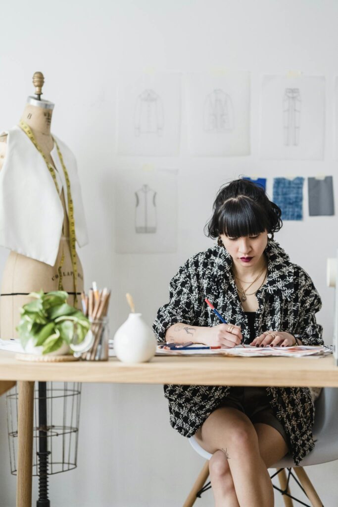 Concentrated Asian female designer sitting at table and drawing sketches against mannequin and light wall with paintings in daytime